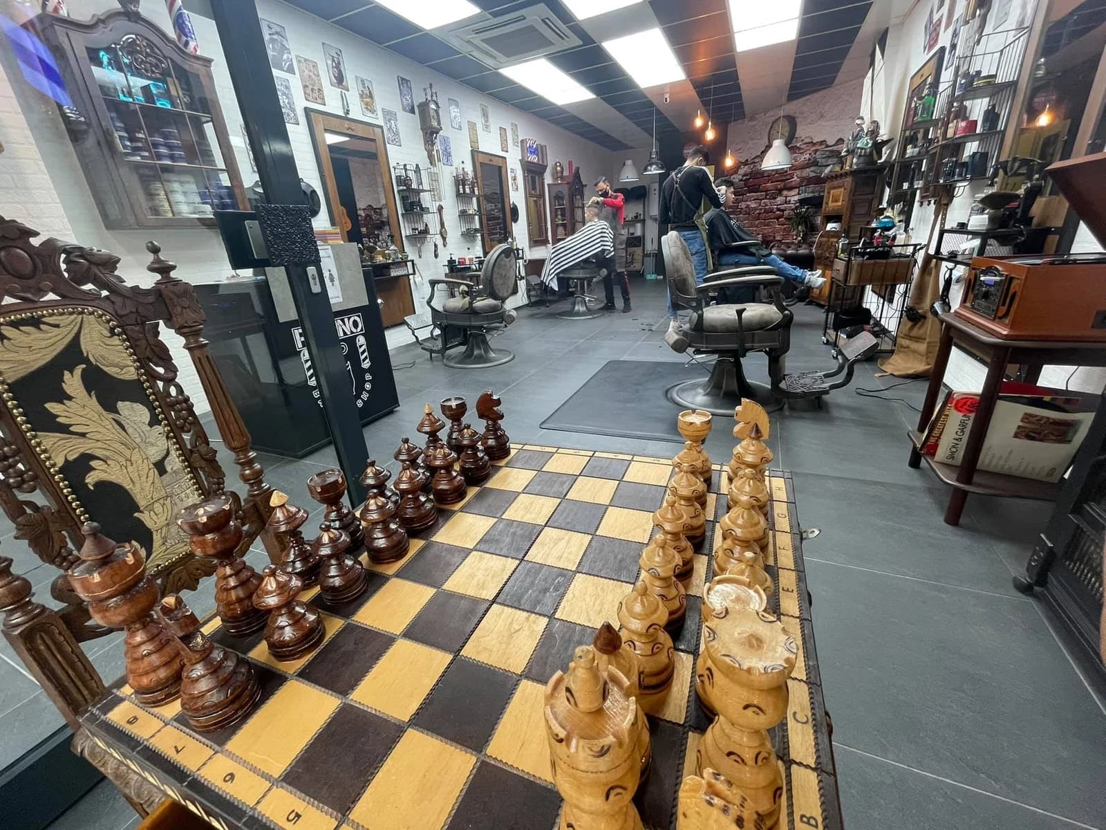 Een houten schaakbord op de voorgrond terwijl de barbers klanten knippen